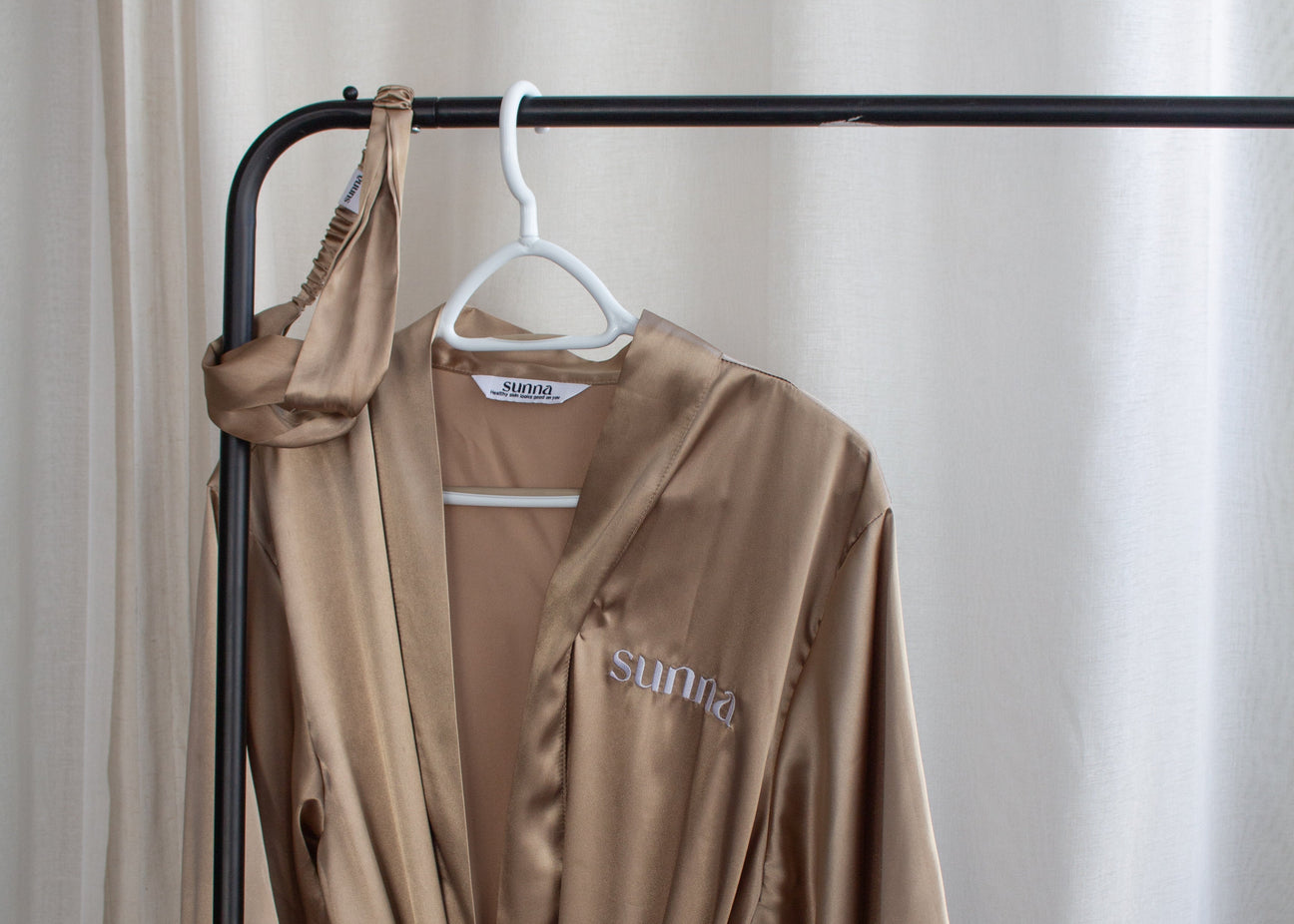 Brown satin robe hanging on a black rack against a white curtain.